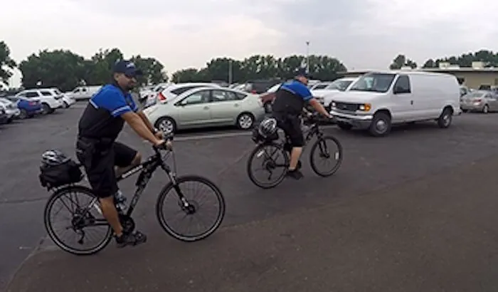 Two Adams County Sheriff Bike Unit officers on bicycles patrolling a parking lot with aparked cars.