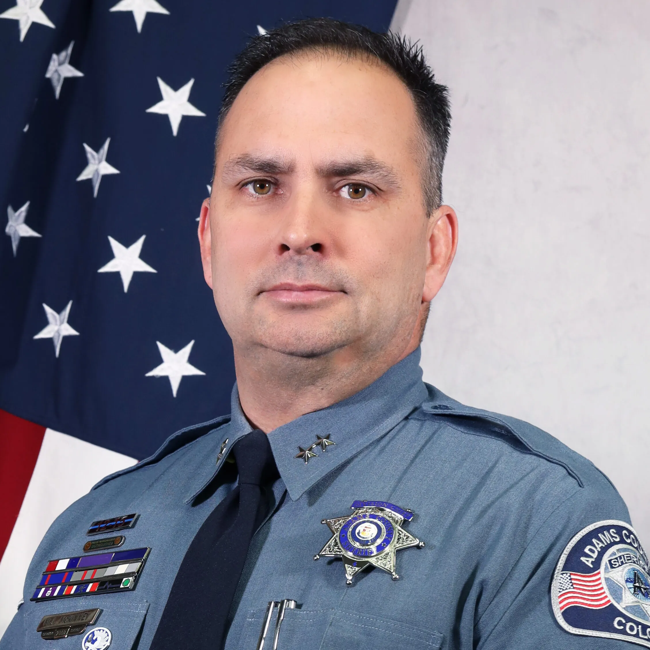 Portrait of Division Chief Mike McKinney in uniform, smiling in front of an American flag. He wears a badge and tie, with the Adams County Sheriff’s Office patch visible on his left sleeve.