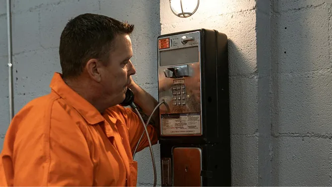 Inmate in orange uniform using a wall-mounted payphone under dim light.