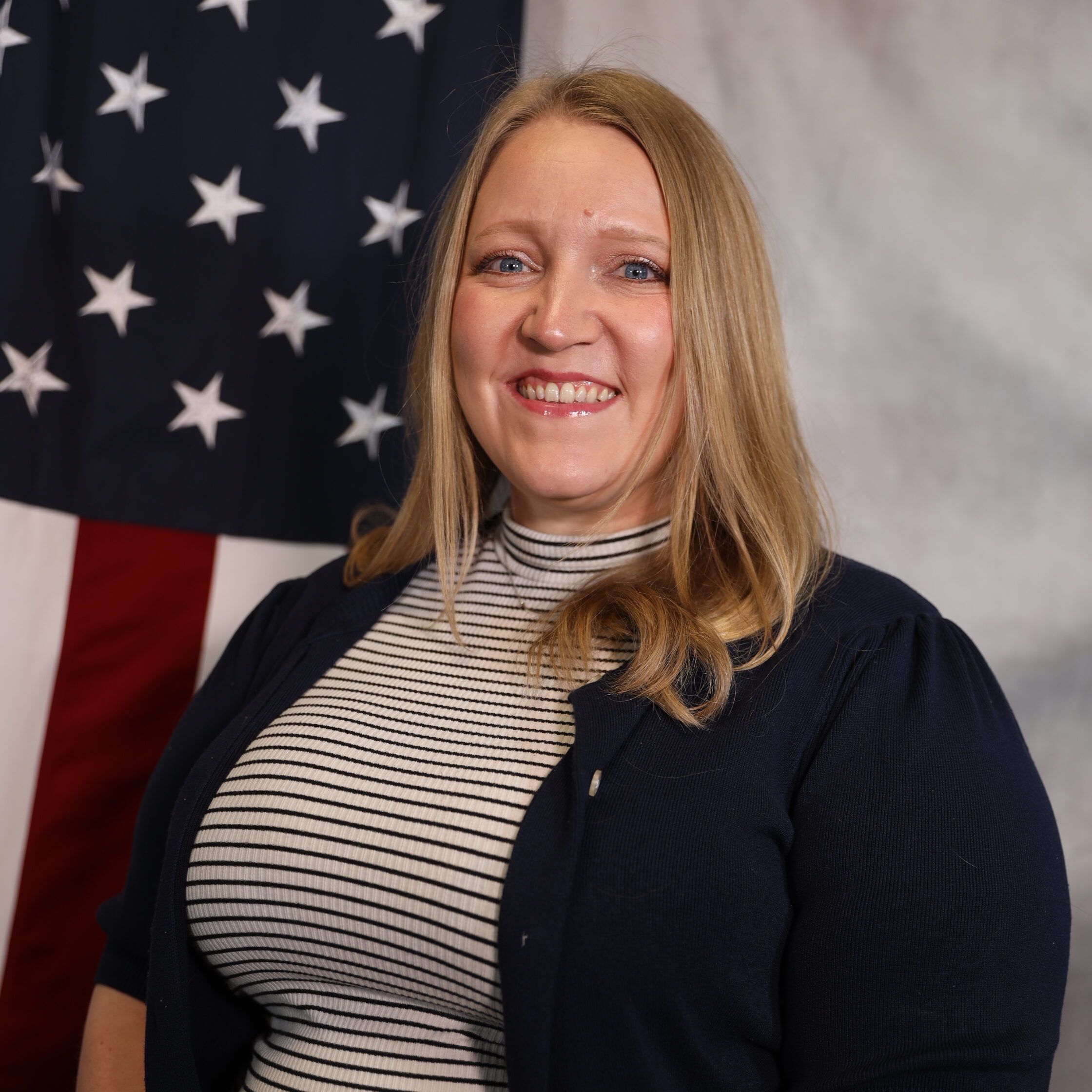Desiree smiling in front of an American flag, wearing a striped top and cardigan.