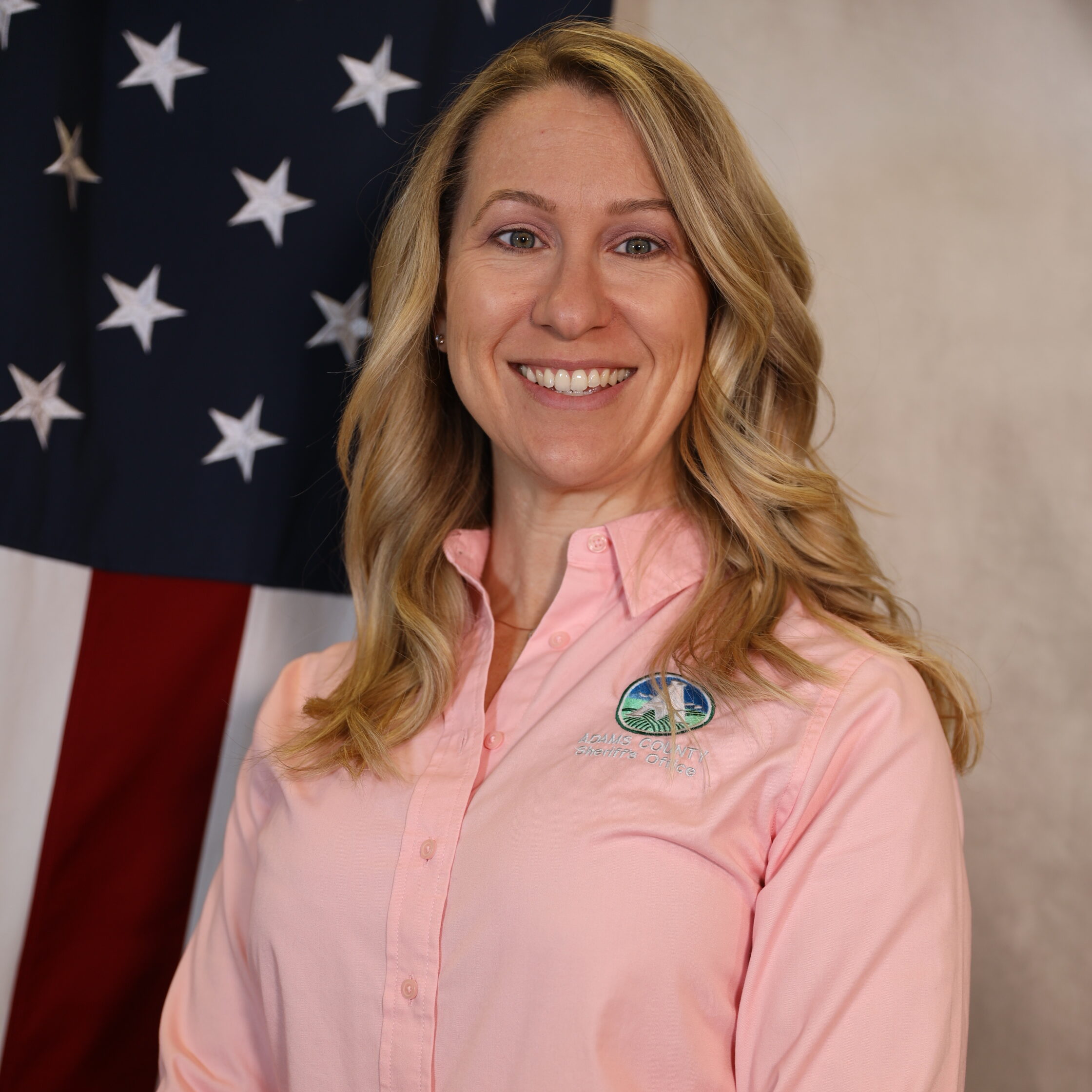 Narverud-Nicole smiling, wearing a pink shirt with a badge, standing in front of an American flag