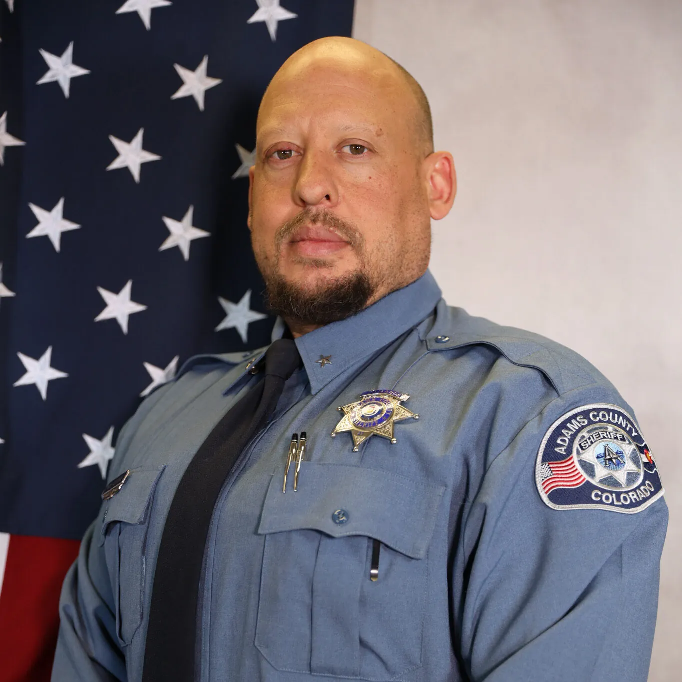 Robbins-Michael-2.jpg: Person in uniform poses confidently before an American flag with visible badge.