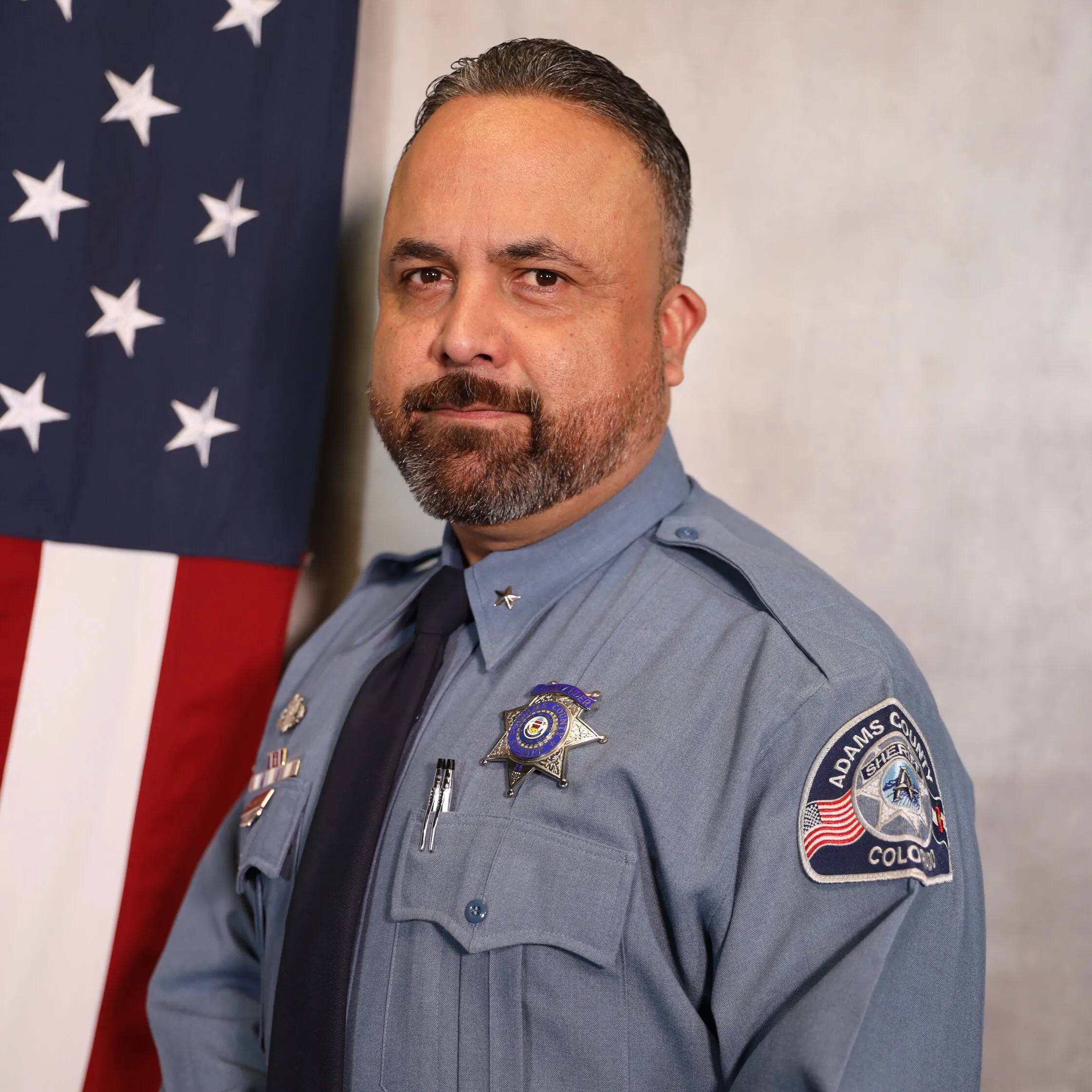 Carrillo-Manuel posing in uniform with U.S. flag background, serious expression, wearing badges on left chest and shoulder.