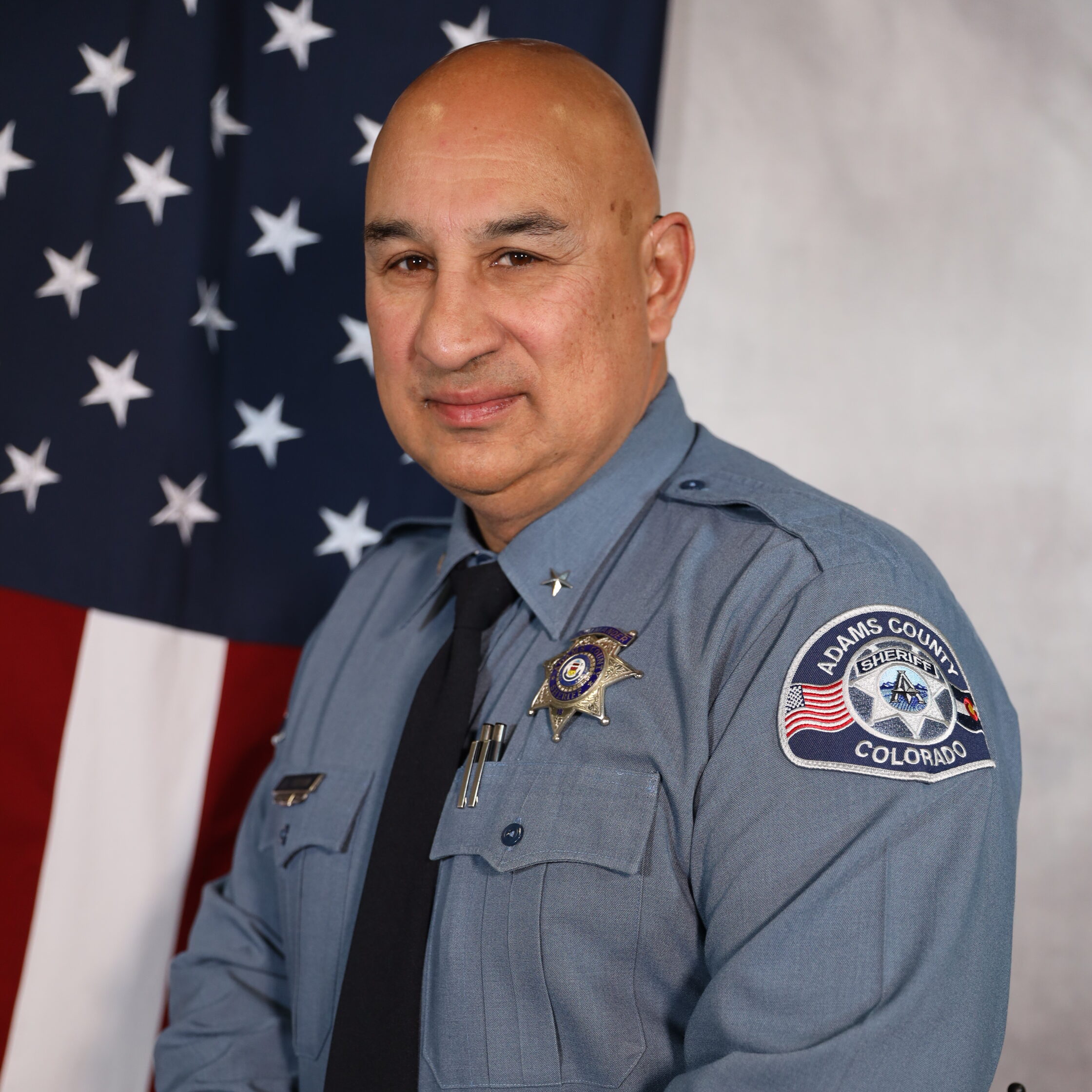 Cordova J.D. in uniform, standing in front of the American flag, Adams County Sheriff's Department patch visible on sleeve.