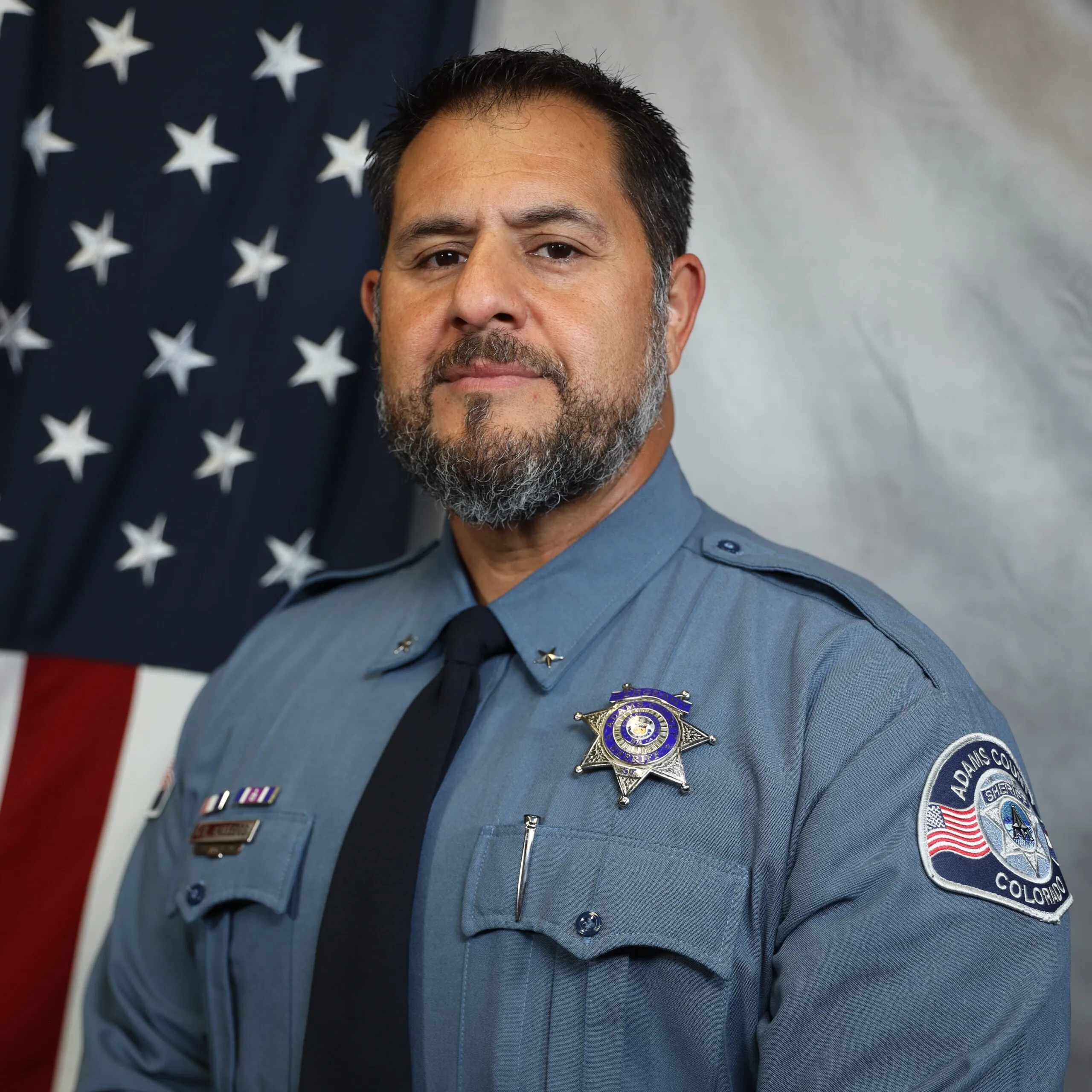 Jason Gallegos, in uniform with badge, standing before U.S. flag