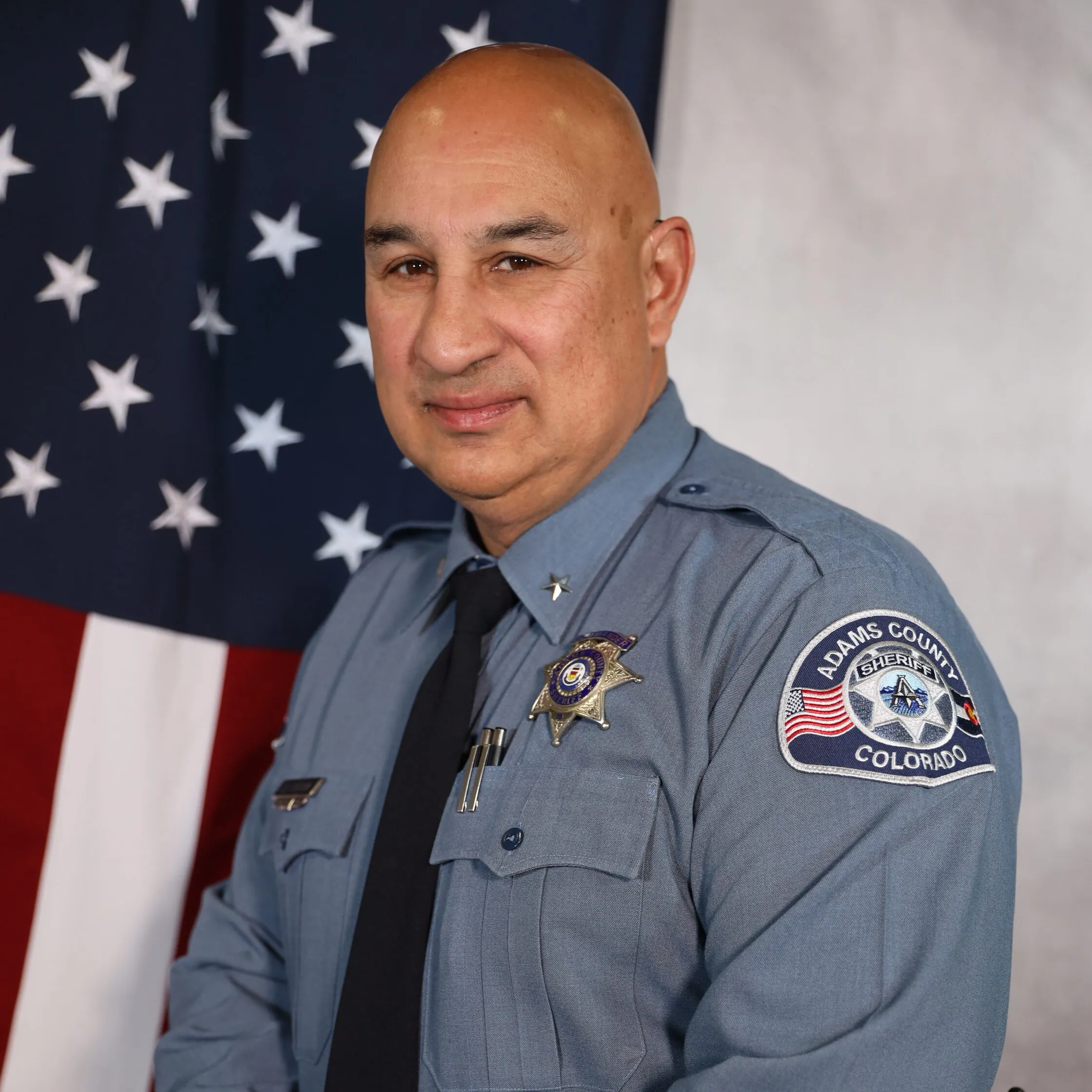 Cordova J.D. in uniform, standing in front of the American flag, Adams County Sheriff's Department patch visible on sleeve.