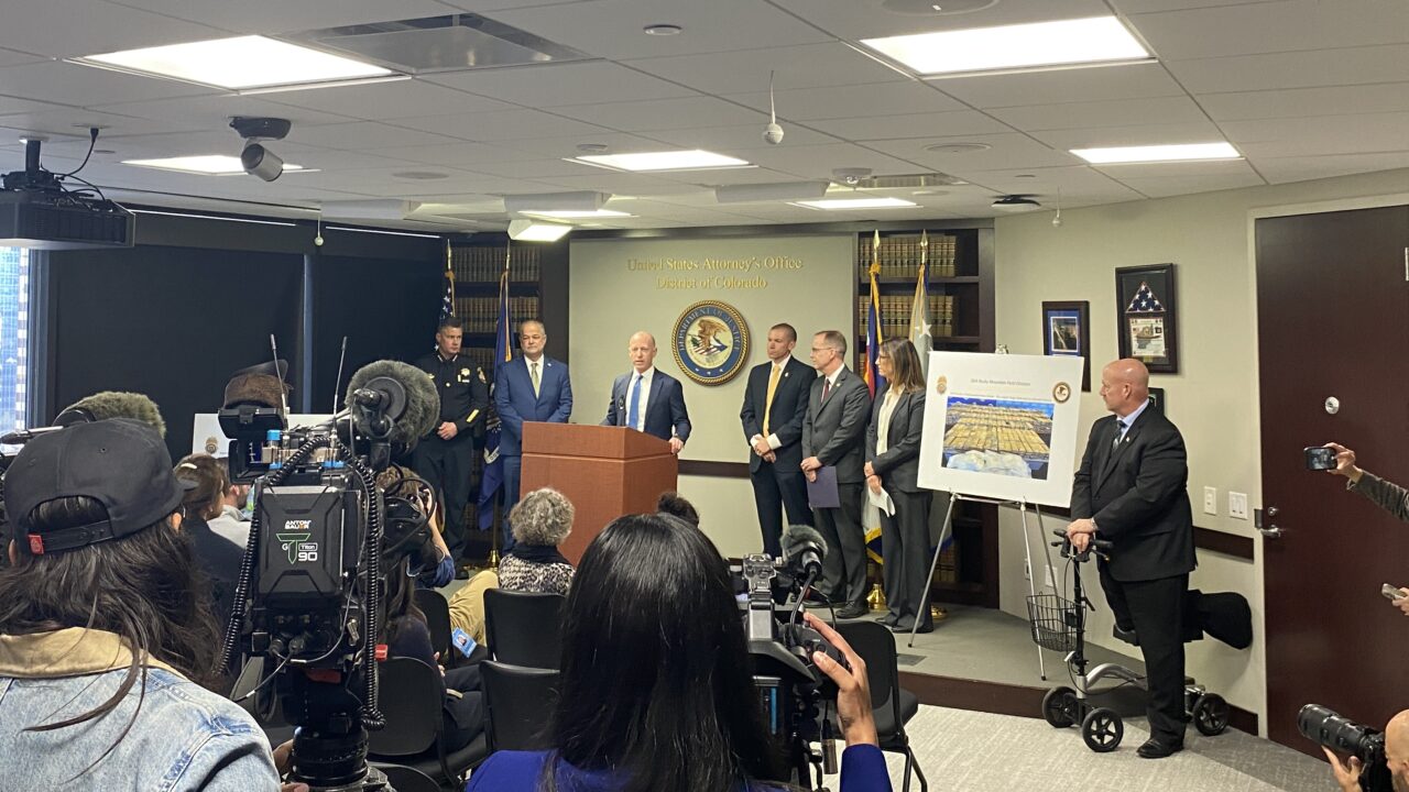 Press conference with multiple cameras, officials at podium, and a large poster.