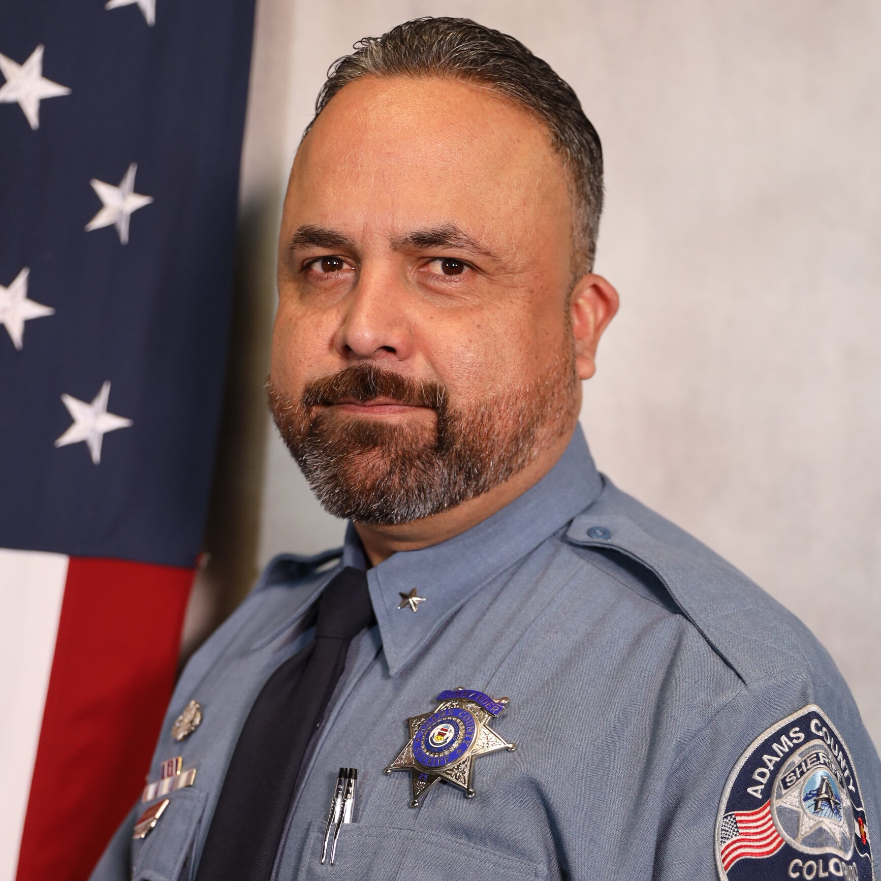 Carrillo-Manuel posing in uniform with U.S. flag background, serious expression, wearing badges on left chest and shoulder.