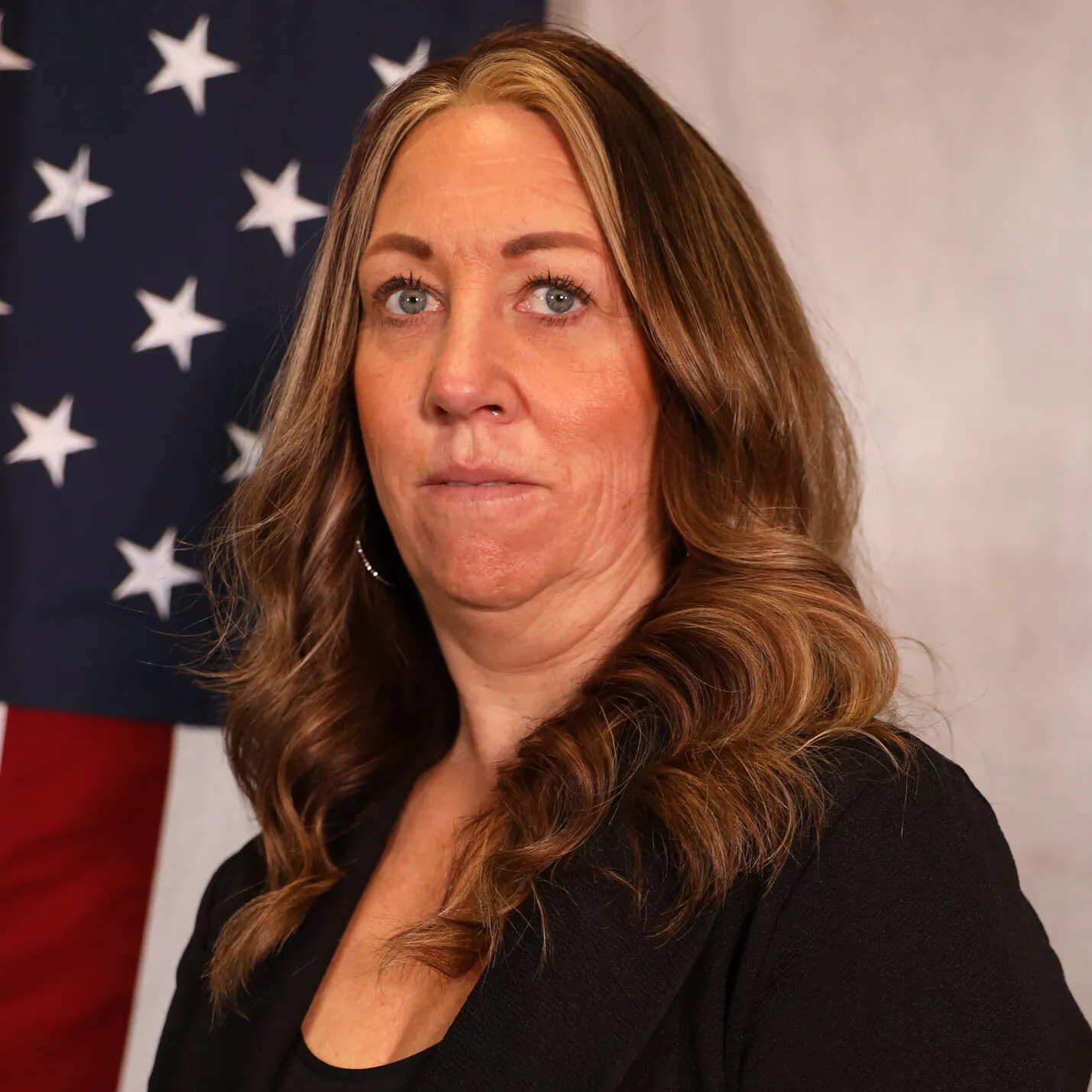Jana standing in front of an American flag, wearing a black blazer, with a serious expression
