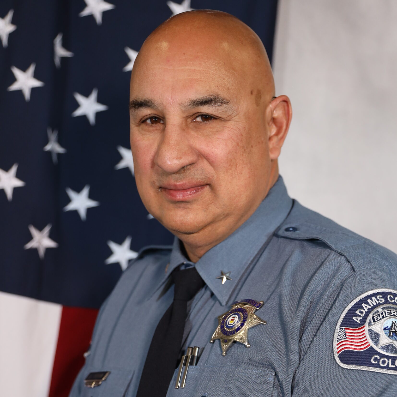 Cordova J.D. in uniform, standing in front of the American flag, Adams County Sheriff's Department patch visible on sleeve.