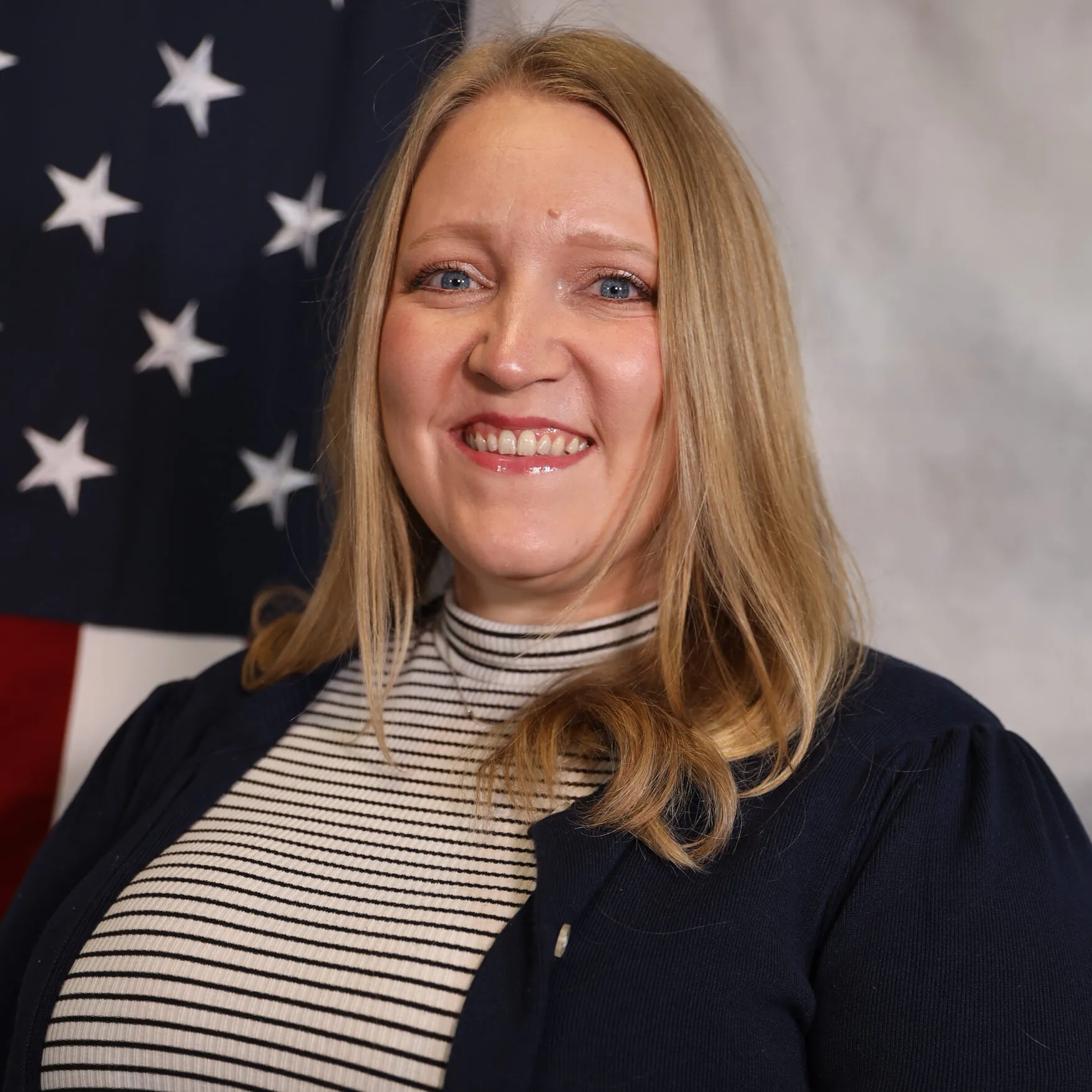Desiree smiling in front of an American flag, wearing a striped top and cardigan.
