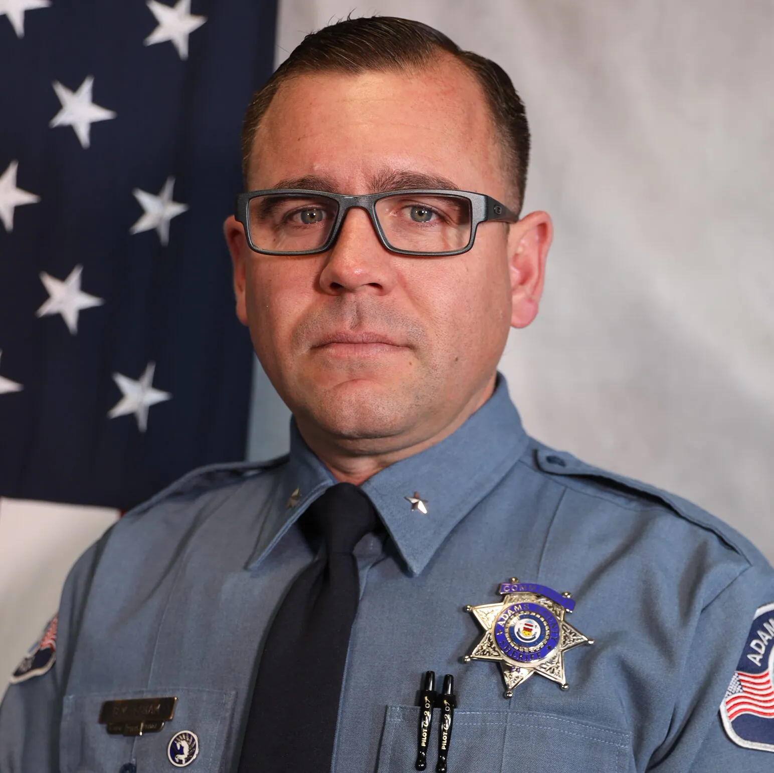 Sheriff wearing a uniform with a badge, standing in front of an American flag