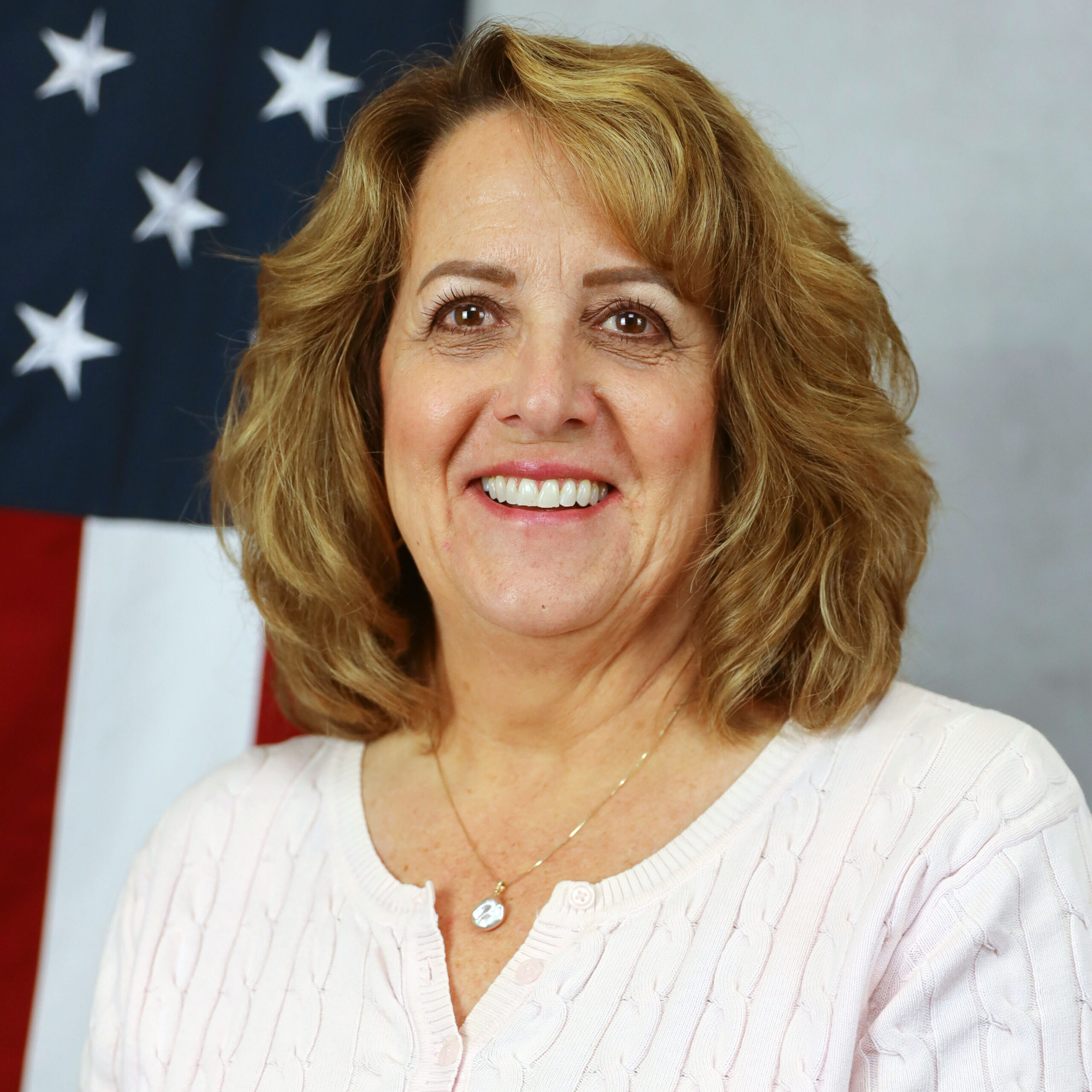 Lori Feola smiling in front of an American flag backdrop, wearing a light pink sweater and necklace