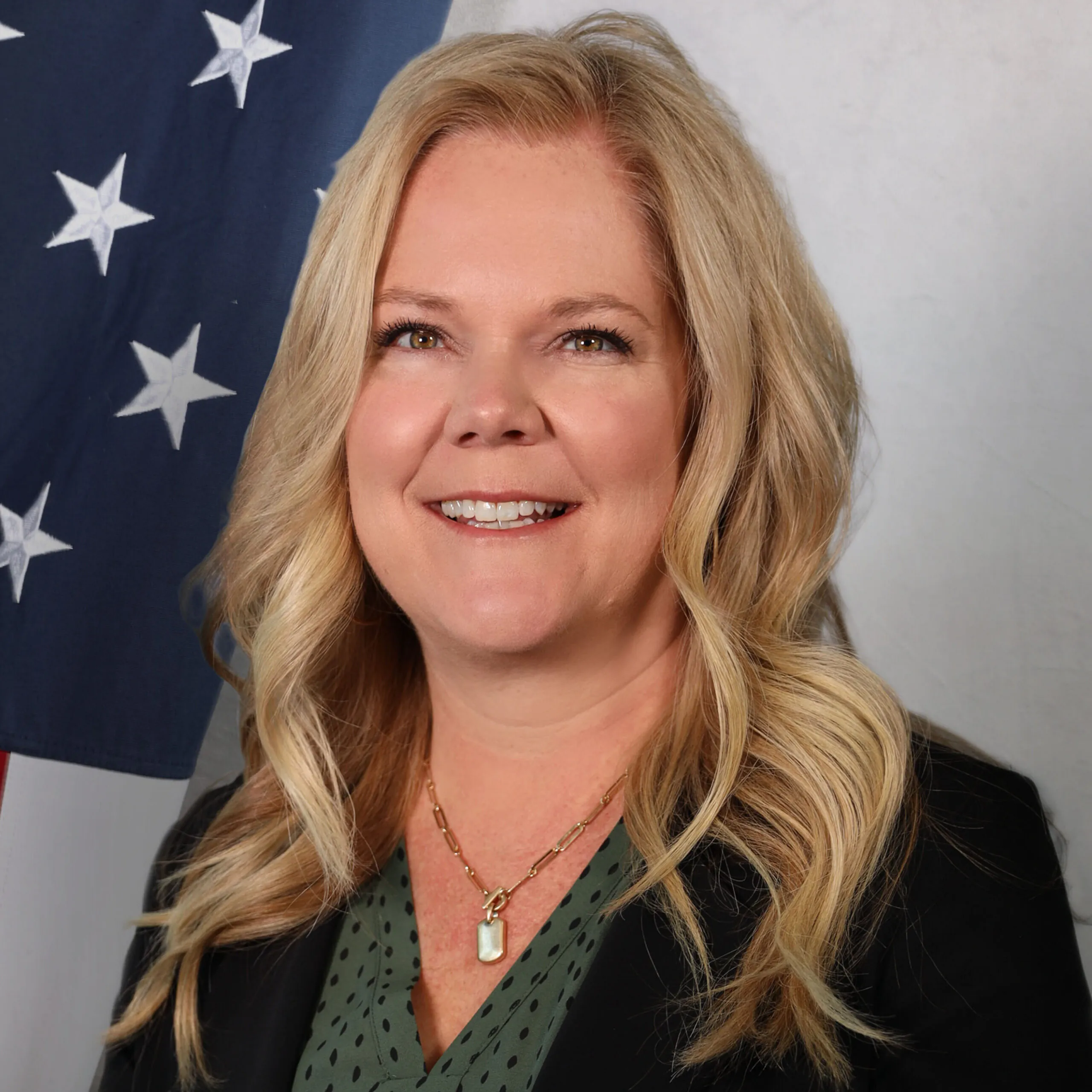 Jennifer Baessler smiles in front of an American flag backdrop, wearing a green blouse and gold necklace