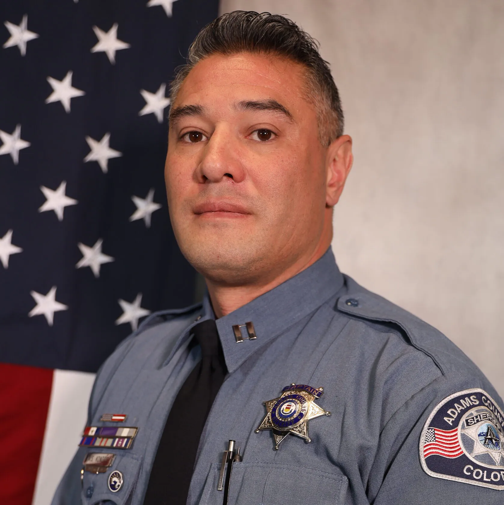 Alex Kondos in an Adams County Sheriff's uniform, standing in front of an American flag