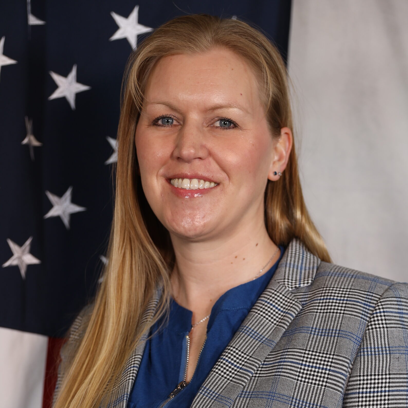 Woman smiling in front of an American flag, wearing a blue shirt and plaid blazer