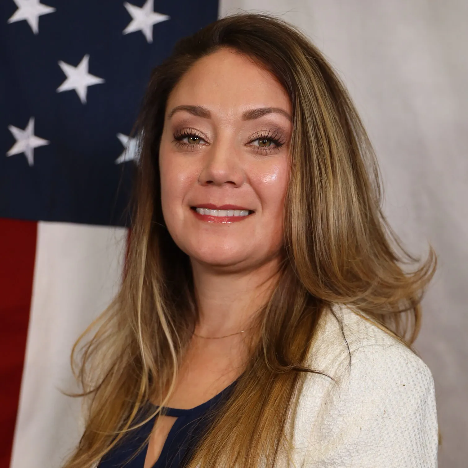 Ashley smiling in front of a U.S. flag backdrop, wearing a white blazer