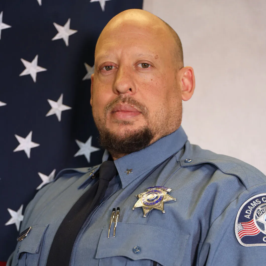 Sheriff in uniform with badge and Adams County patch, standing in front of a star-patterned flag background